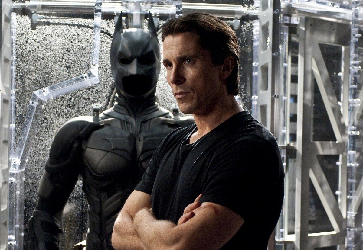 : Christian Bale menggunakan kaos hitam dan terdapat kostum Batman di belakangnya  